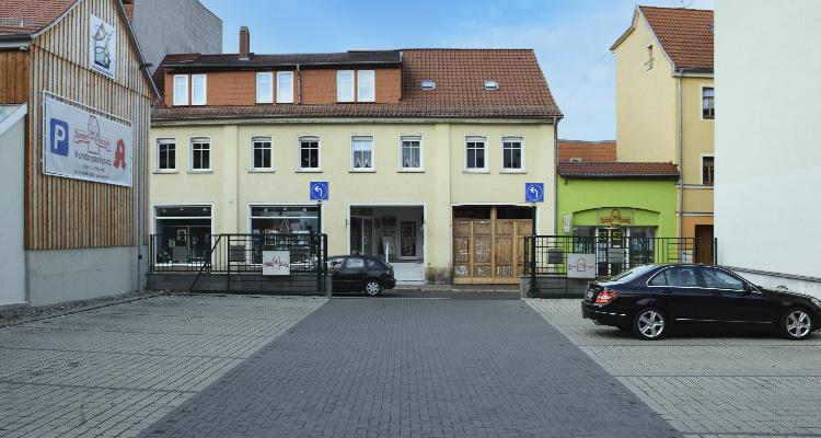 Foto der Parkplatzfläche der Apotheke am Darrplatz. Im Vordergrund steht ein geparktes Auto, rechts im Hintergrund sieht man die Fassade der Apotheke.